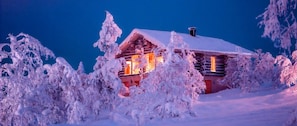 Une cabane en rondins dont le toit et les fenêtres sont recouverts de neige, entourée d’arbres chargés de neige.