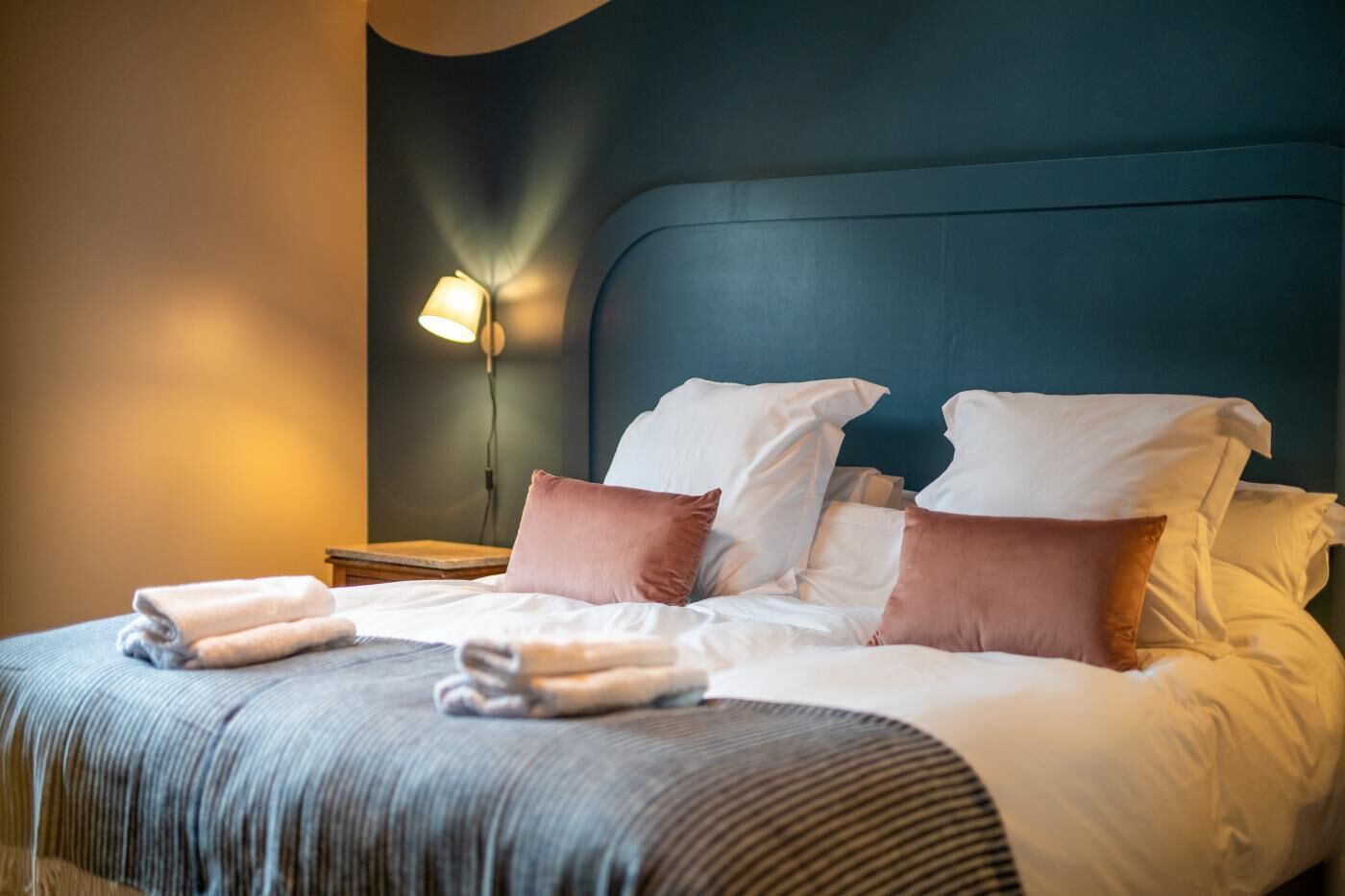 Cama bem arrumada com travesseiros e cobertor, luminária de cabeceira e cabeceira escura.