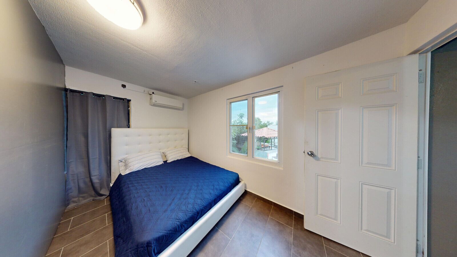 A bedroom with a bed, a window, an air conditioning unit, and a door.
