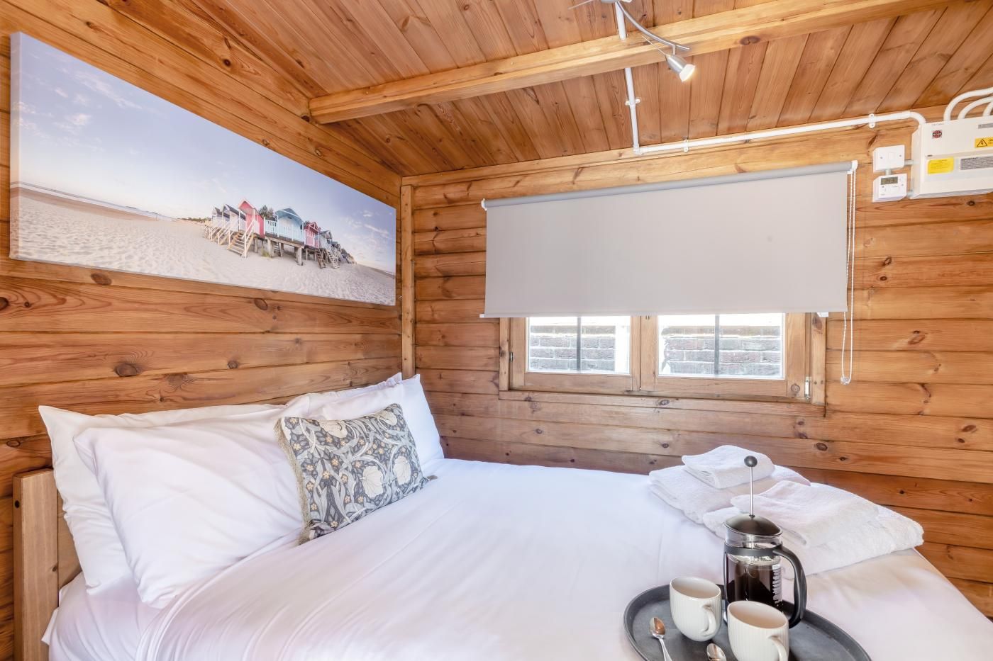 A wooden cabin room with a bed, a painting of a beach scene, a window with a view, and a heater on the wall.