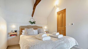 Ein ordentlich bezogenes Bett mit weißen Leinen und flauschigen Kissen, ein Nachttisch mit Lampe und eine Holztür.