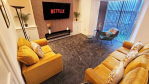 A living room with a flat-screen TV mounted on a wall, two sofas with cushions, a small side table, and a floor lamp. - 4 Bed townhouse Swansea city centre (Swansea)