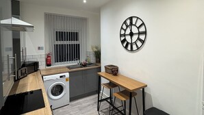 A compact kitchen with a washing machine, a wooden table with stools, and a wall-mounted clock. - 4 Bed townhouse Swansea city centre (Swansea)
