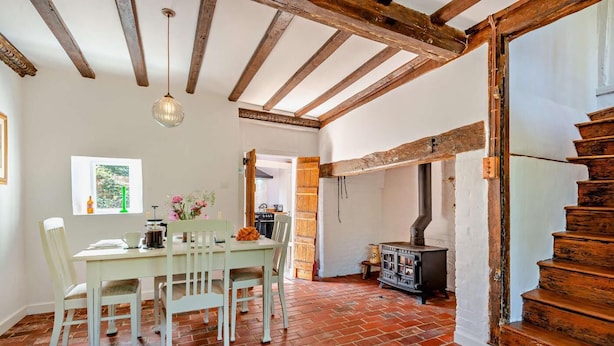 A rustic interior with exposed wooden beams, a brick floor, a dining area with a table and chairs, a wooden staircase, and a wood-burning stove. - 3 Bed in Benenden (oc-cb671) (Benenden)