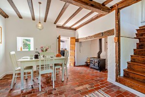A rustic interior with exposed wooden beams, a brick floor, a dining area with a table and chairs, a wooden staircase, and a wood-burning stove. - 3 Bed in Benenden (oc-cb671) (Benenden)