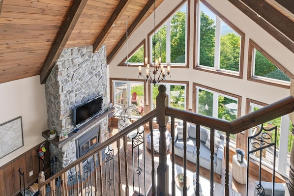 A spacious living area with a stone fireplace, large windows, and a wooden staircase.