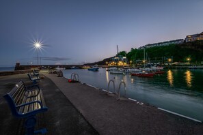 Une promenade en bord de mer avec des bancs, un port tranquille où sont amarrés des bateaux, et un bâtiment illuminé d’un côté.