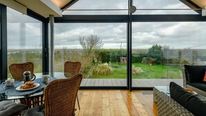 A modern room with a large glass wall, a dining table with chairs, and a sofa. - 2 Bed in Morston (oc-2382) (Morston)