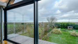 A modern room with large glass windows overlooking a lush garden and a distant landscape. - 2 Bed in Morston (oc-2382) (Morston)