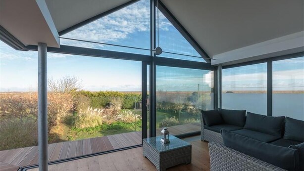 A modern room with large glass windows, a wooden floor, a grey sofa, and a small table with a lantern. - 2 Bed in Morston (oc-2382) (Morston)