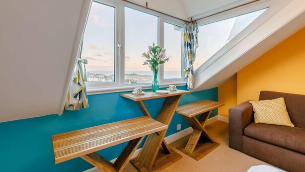 Ein Zimmer mit schrägem Dach, einem fenster mit Vorhängen, einem Holztisch, einem braunen Sofa und einer Vase mit Blumen.