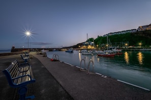 A waterfront promenade with benches, a calm harbor with boats, and a building with lights on one side. - 4 Bed in Saundersfoot (oc-pw9285) (Saundersfoot)