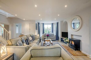 A spacious living room with a sofa set, a fireplace, a television, and a large window with curtains. - 6 Bed in Bacton (oc-2316) (Bacton)