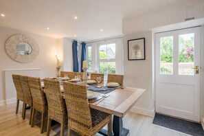 A dining area with a round mirror, wicker chairs, a white door, and a window with a view of greenery. - 6 Bed in Bacton (oc-2316) (Bacton)