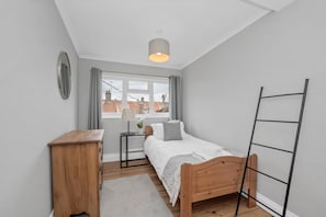 A bedroom with a bed, a desk, a radiator, a window with curtains, and a view of buildings outside. - 4 Bed in Southwold (oc-stjam) (Southwold)