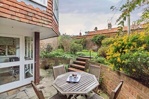 A patio with a table and chairs, a brick wall, and a garden with yellow flowers. - 4 Bed in Southwold (oc-stjam) (Southwold)