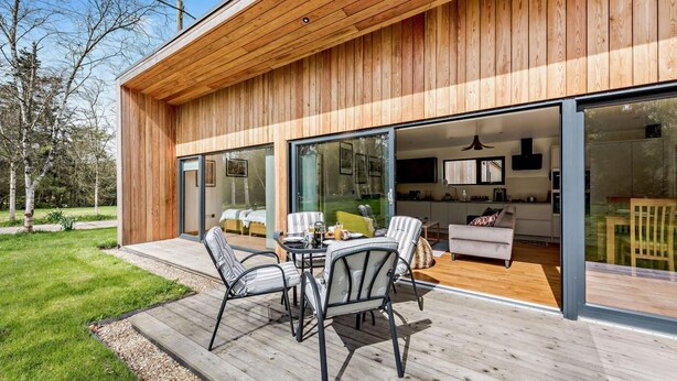 A modern wooden house with a deck, sliding glass doors leading to a living area with a sofa and a dining table.