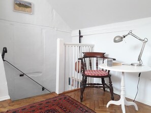 Habitación de esquina con un radiador blanco, una silla de madera con cojín rojo, una mesita redonda con lámpara y una alfombra estampada en el suelo.