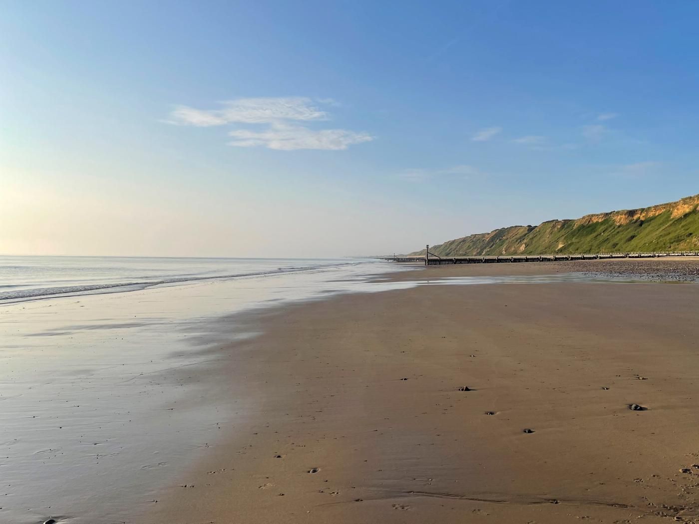 Une plage avec une jetée en bois qui s’avance dans l’eau, un ciel dégagé et une colline lointaine.