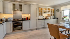 A modern kitchen with white cabinetry, a dining area with a wooden table and chairs, and a window with a view. - 3 Bed in Southwold (oc-s27203) (Reydon)