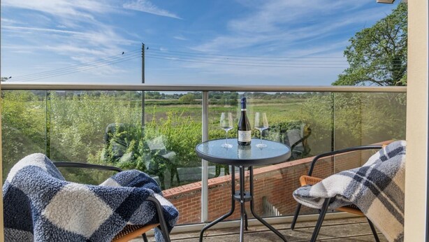A balcony with a glass table, two chairs, and a bottle of wine. - 3 Bed in Southwold (oc-s27203) (Reydon)