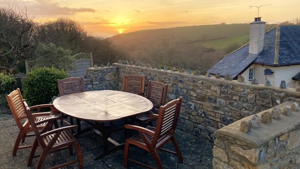 A stone wall patio with a round wooden table and chairs, overlooking a rural landscape and a house with a chimney. - 4 Bed in Tenby (oc-vv27177) (Manorbier)