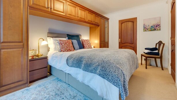 Un dormitorio con una cama, armarios de madera, una mesita de noche, una silla y un cuadro en la pared.