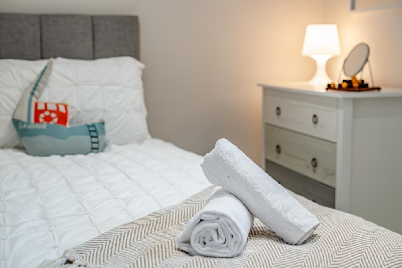 A neatly made bed with a white comforter, a rolled white towel, and a grey headboard.