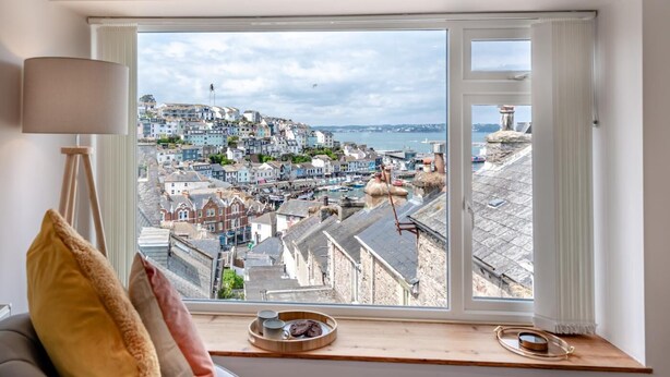A room with a large window overlooking a coastal town, a lamp, and a tray with a mug and cookies on the windowsill. - 2 Bed in Brixham (oc-s27146) (Brixham)