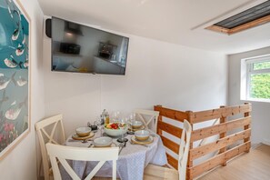 Un coin repas avec une table dressée pour quatre personnes, un téléviseur fixé au mur au-dessus, et une fenêtre donnant sur un paysage verdoyant.