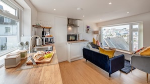 A modern kitchen with white cabinets, a dining area with a dark blue sofa, and a view of the city through large windows. - 2 Bed in Brixham (oc-s27146) (Brixham)