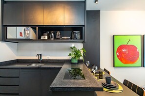 Eine moderne Küche mit integriertem Mikrowellenherd, Kaffeemaschine und Toaster. Es gibt ein Spülbecken mit Armatur, eine Arbeitsfläche mit Pflanze und einen Essbereich mit Tisch für zwei Personen.