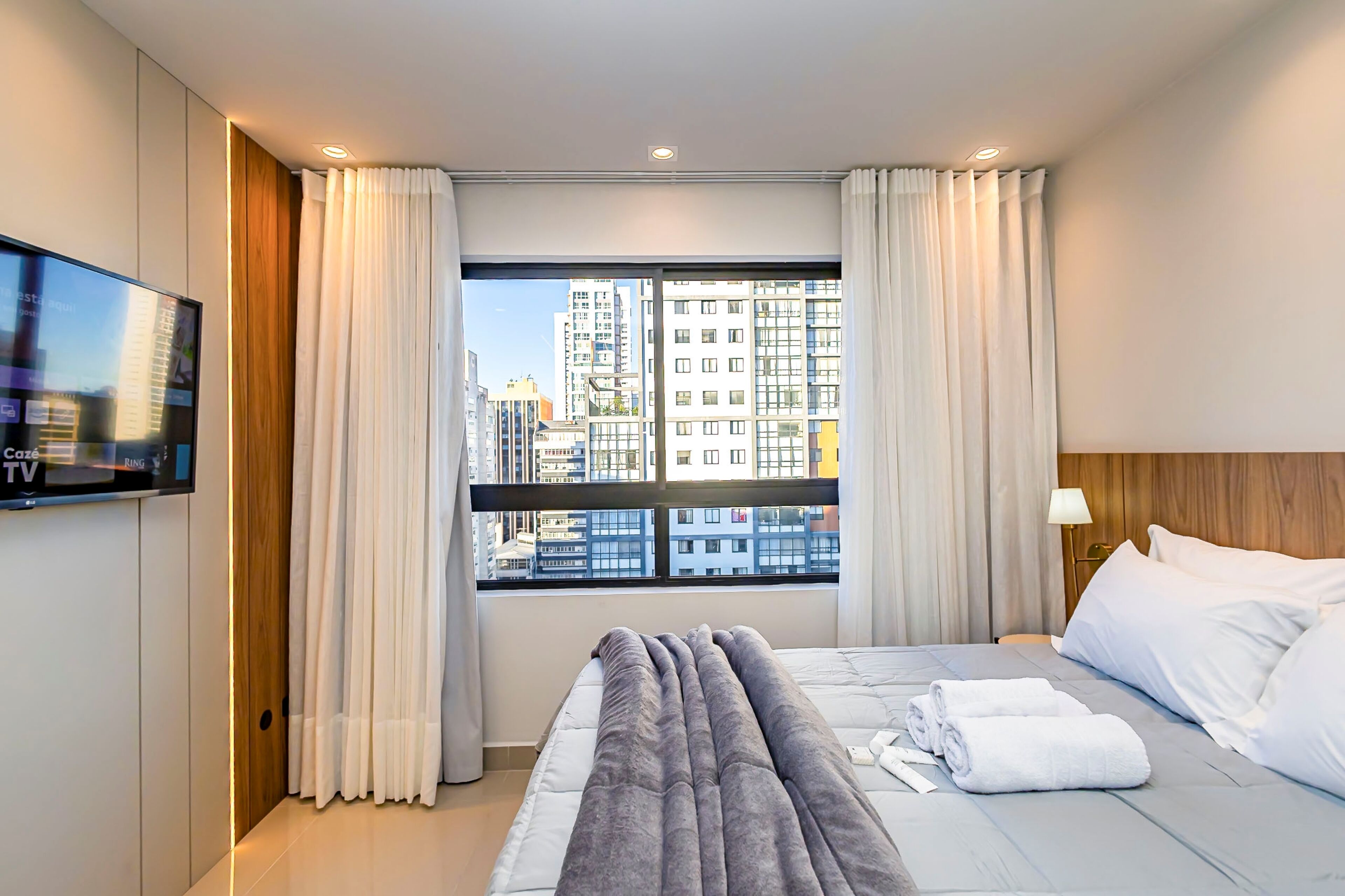 Una habitación de hotel con cama, televisor, ventana con cortinas y vista a la ciudad.