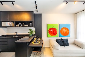 Eine moderne Küche mit einer zentralen Kochinsel, einem Essbereich mit einer Couch und zwei Wandbildern mit Obstmotiven.