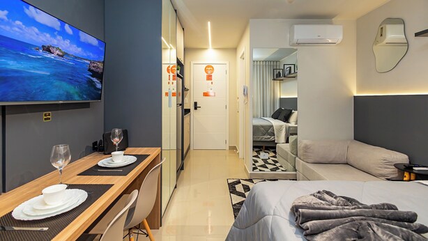 A modern hotel room with a TV, a dining area with a table set for two, a bed with a gray blanket, a sofa, and an air conditioning unit. - MUV Residence (Curitiba)