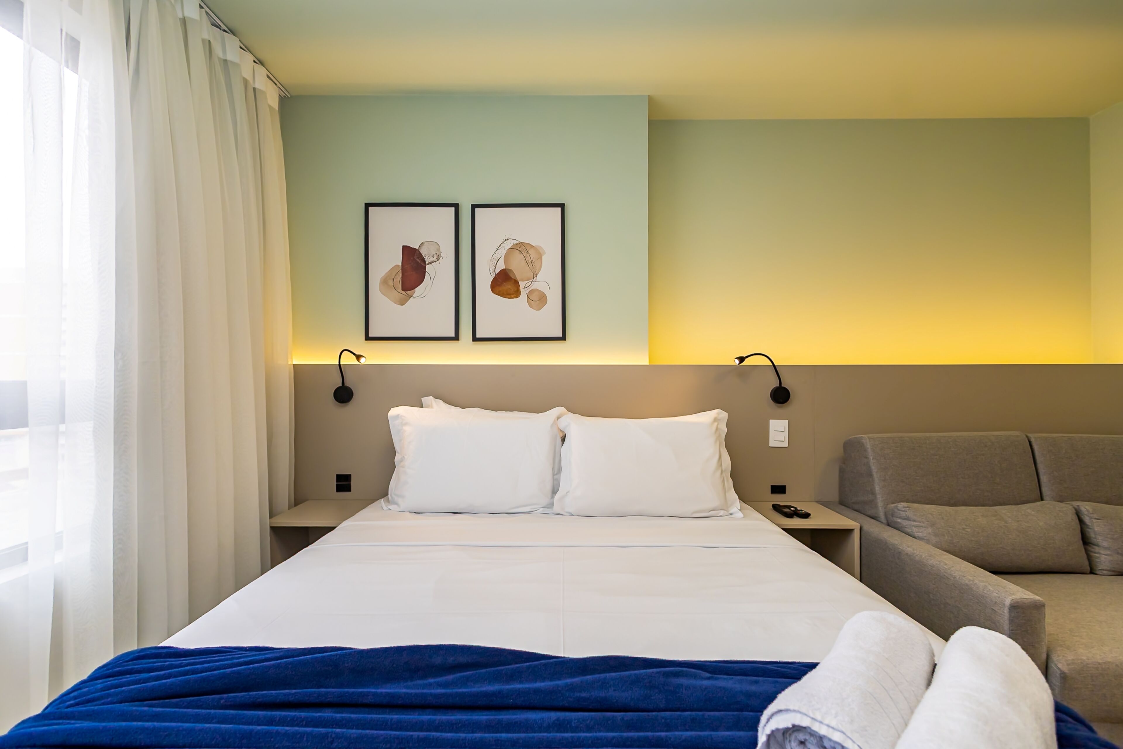 A hotel room with a bed, a sofa, two framed artworks on the wall, and a nightstand with a lamp.