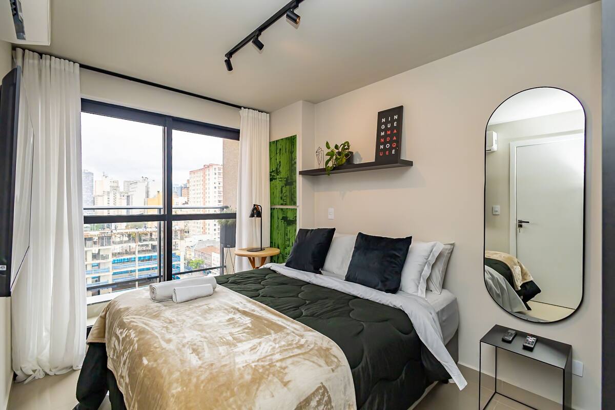 Una habitació d'hotel amb un llit, una finestra gran amb cortines, una taula de nit amb una làmpada, un mirall i una decoració de paret verda.