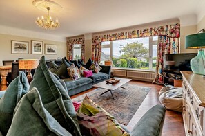 A living room with a sofa, a coffee table, and a television. There are curtains and a chandelier. - 10 Bed in Llanrhidian (oc-s27225) (Pen-clawdd)