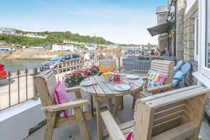 A wooden deck with a table set for dining, overlooking a harbor with boats and buildings in the background. - 3 Bed in Porthleven (oc-1seav) (Porthleven)