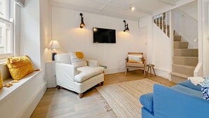 Uma sala de estar com um sofá branco, uma poltrona azul, uma cadeira de madeira, uma televisão fixada na parede e uma escada.