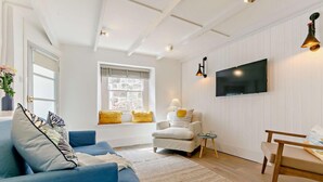Uma sala de estar com um sofá azul, uma poltrona branca, uma mesa de madeira e uma TV fixada na parede.
