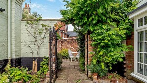 A narrow garden pathway leading to a white chair, surrounded by potted plants and a brick wall. - 2 Bed in Yoxford (oc-c27253) (Wickham Market)