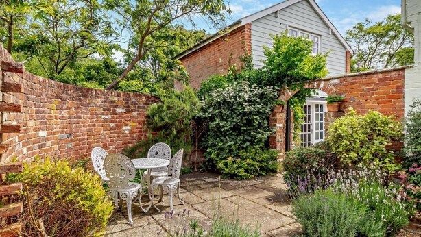 A brick house with a white door, surrounded by a garden with various plants and a patio area with a table and chairs. - 1 Bed in Yoxford (oc-c27254) (Wickham Market)
