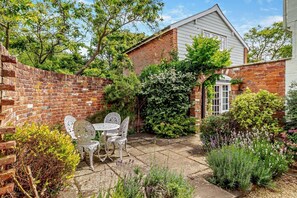 A brick house with a white door, surrounded by a garden with various plants and a patio area with a table and chairs. - 1 Bed in Yoxford (oc-c27254) (Wickham Market)