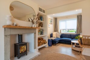 A living room with a fireplace, a blue sofa, a round table, and a large window with curtains. - 3 Bed in Blythburgh (oc-h27263) (Blythburgh)