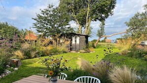 Une terrasse en bois avec une table et des chaises, entourée d’un jardin où poussent différentes plantes et adossée à une petite construction en bois.