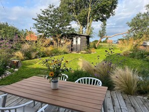 A wooden deck with a table and chairs, surrounded by a garden with various plants and a small wooden structure. - 3 Bed in Blythburgh (oc-h27263) (Blythburgh)