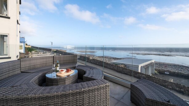 A balcony with wicker furniture, a table with a bottle and bowl, and a view of the sea and rocky shore. - 4 Bed in Amroth (oc-g27016) (Amroth)