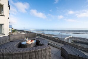 Een balkon met rieten meubilair, een tafel met fles en schaal, en uitzicht op zee en een rotsachtige kust.