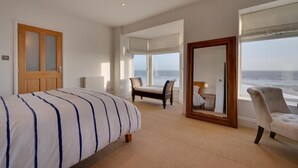 A bedroom with a bed, a chair, a window with a view of the ocean, and a mirror reflecting the room. - 4 Bed in Amroth (oc-g27016) (Amroth)